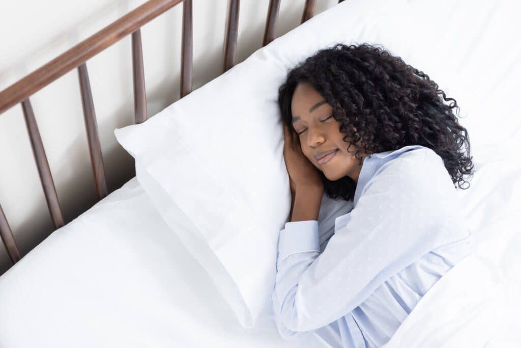 Une femme place ses mains sous sa tête sur un oreiller blanc et moelleux, couverte de ses draps en satin de coton.