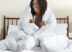 Une femme enroule confortablement la couette alternative en duvet Juno autour de son corps tout en étant assise sur un matelas Juno.