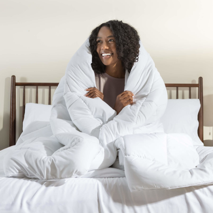 Une femme enroule confortablement la couette alternative en duvet Juno autour de son corps tout en étant assise sur un matelas Juno.