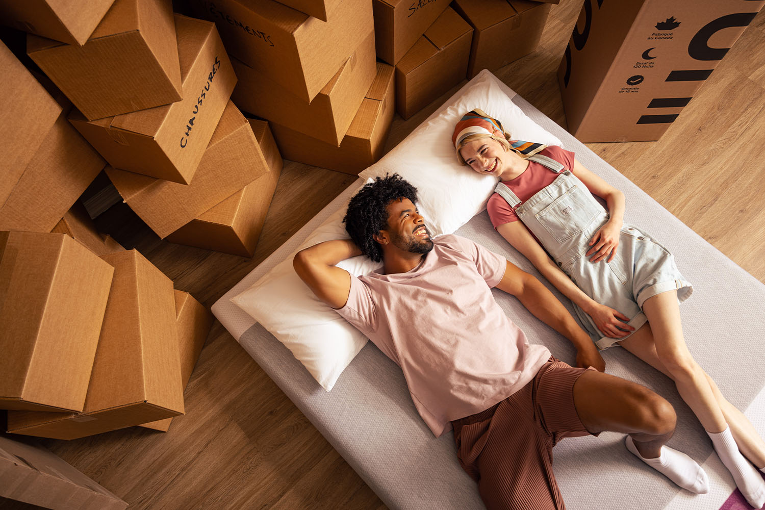 A young man and woman lying down happily on a Juno mattress, surrounded by moving boxes