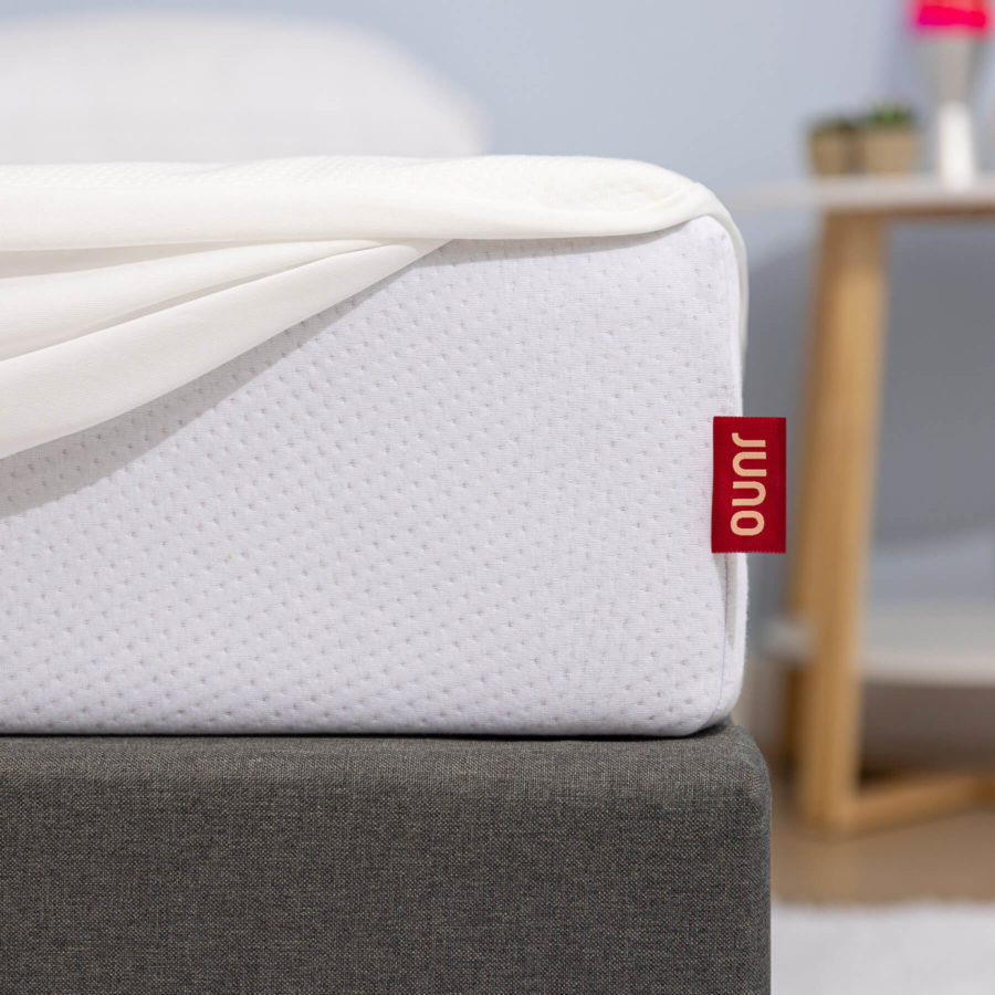The corner of a bed set with a Juno mattress features a mattress protector that is slightly pulled back, revealing a bright red and yellow Juno tag.