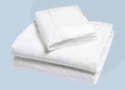 An angled view of a Juno top sheet, fitted sheet, and pillow cover sit neatly folded against a light blue background.