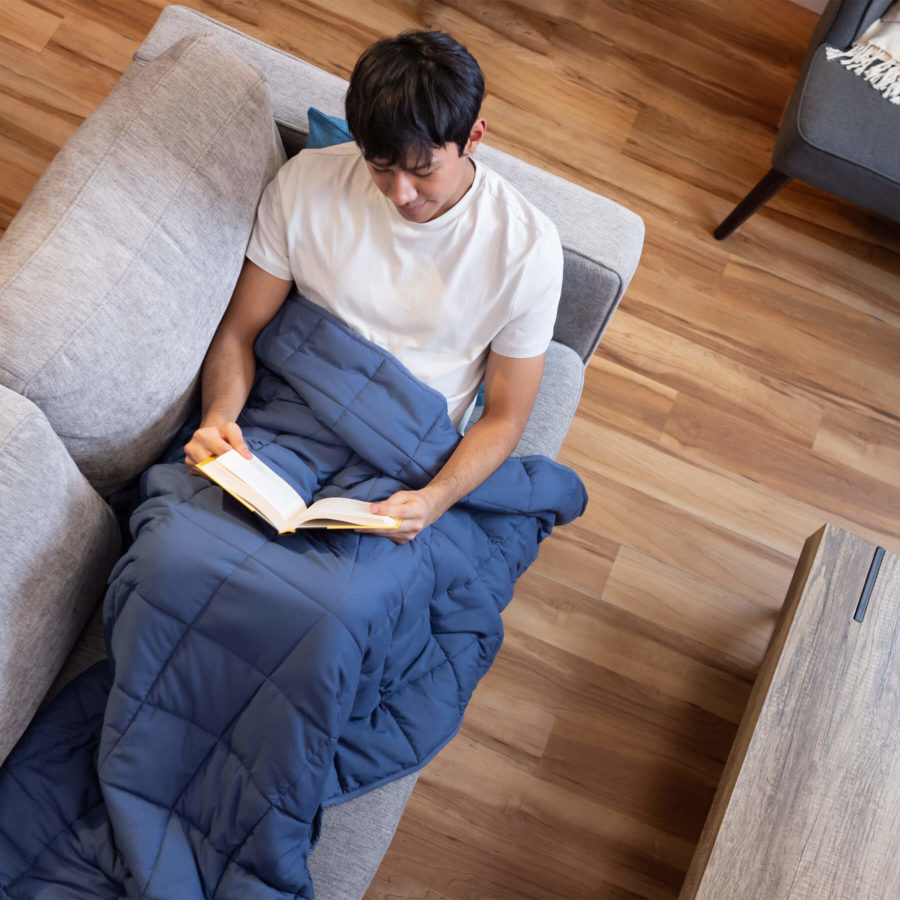 Un homme lit un livre sur le canapé, avec une couverture lestée Juno drapée sur le bas de son corps.