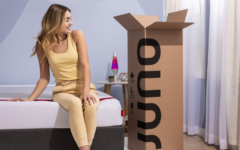 Woman smiling and sitting on a Juno mattress, looking at the opened box